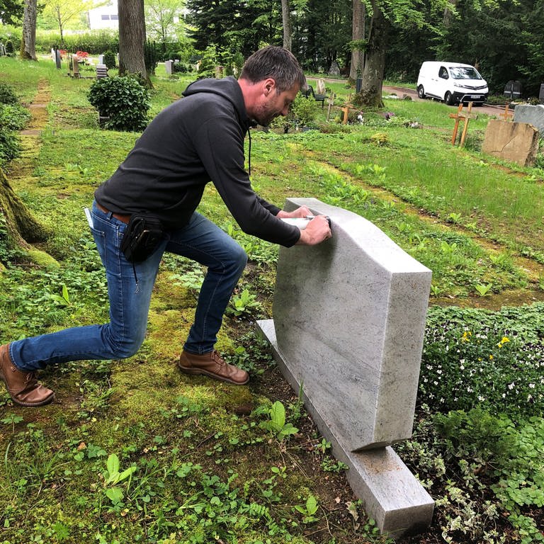 Ein Mann drückt mit Kraft gegen einen hellgrauen Grabstein auf einem Friedhof in Aalen, im Hintergrund Bäume, Gräber und Wiese. (Foto: SWR, Frank Polifke)
